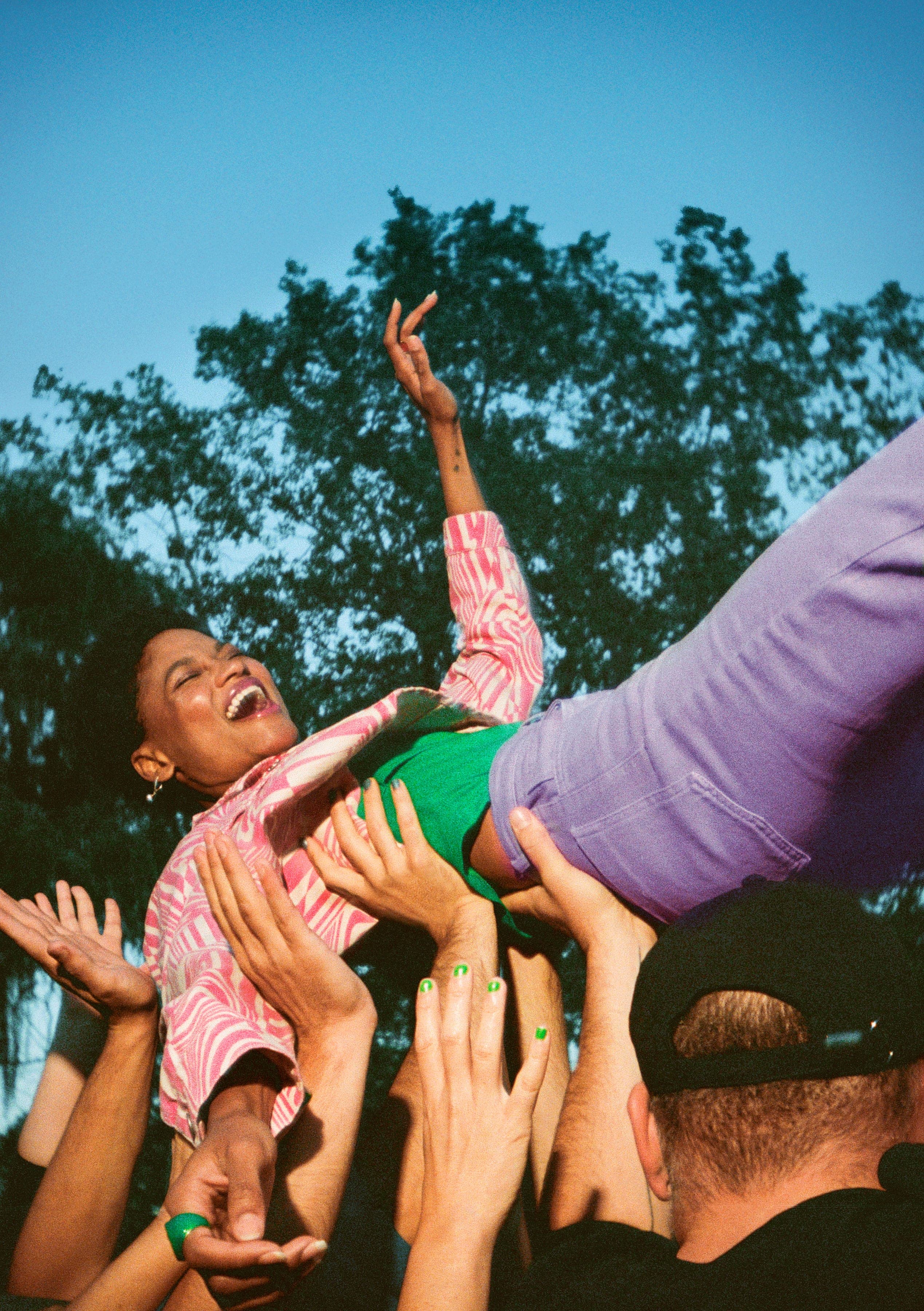 A woman laughing, held up in celebration by a crowd of hands, outdoors under trees.